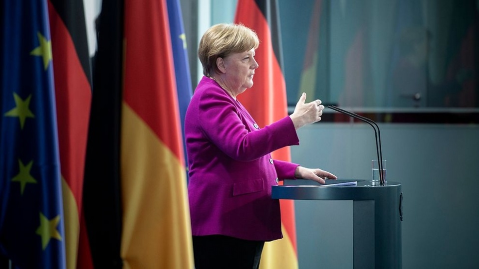 Angela Merkel steht an einem Rednerpult vor einer großen deutschen Flagge, spricht und gestikuliert mit den Händen.