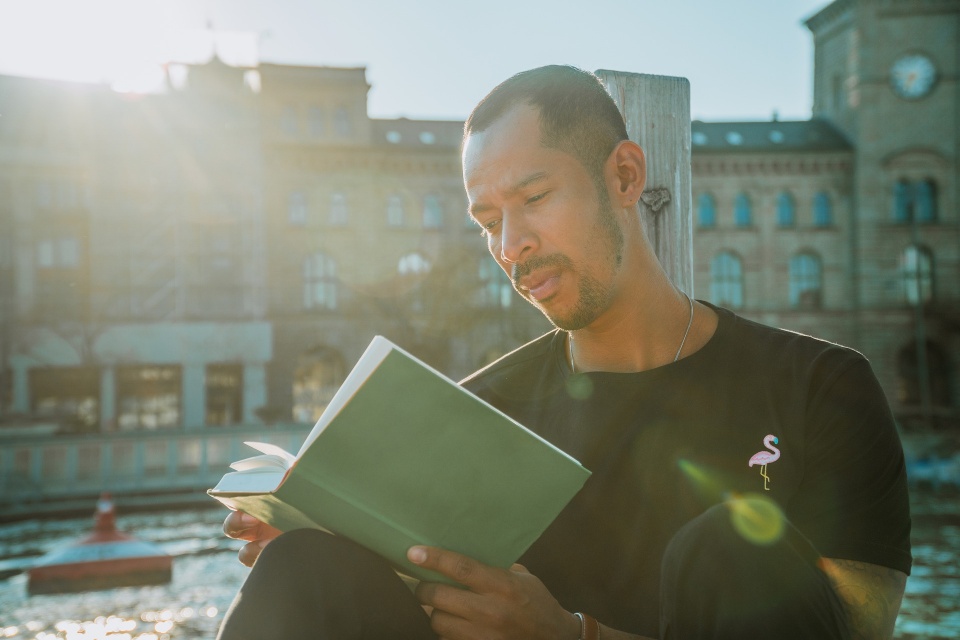 Ein asiatischer Mann liest in einem Buch. Im Hintergrund sieht man eine Häuserkette.