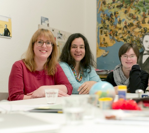 3 Frauen, eine davon mit Down-Syndrom, sitzen nebeneinander an einem Tisch und lächeln.