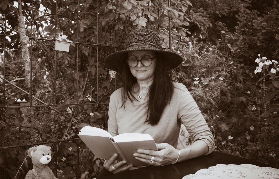 Ein Frau mit langen Haaren, Hut und Brille sitzt im Freien und liest aus einem Buch vor.