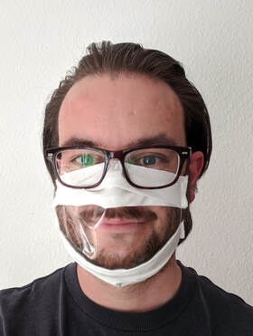 Ein Mann mit Brille und einer Mund Nase Abdeckung mit einem transparenten Mittelteil. Man sieht den Mund.